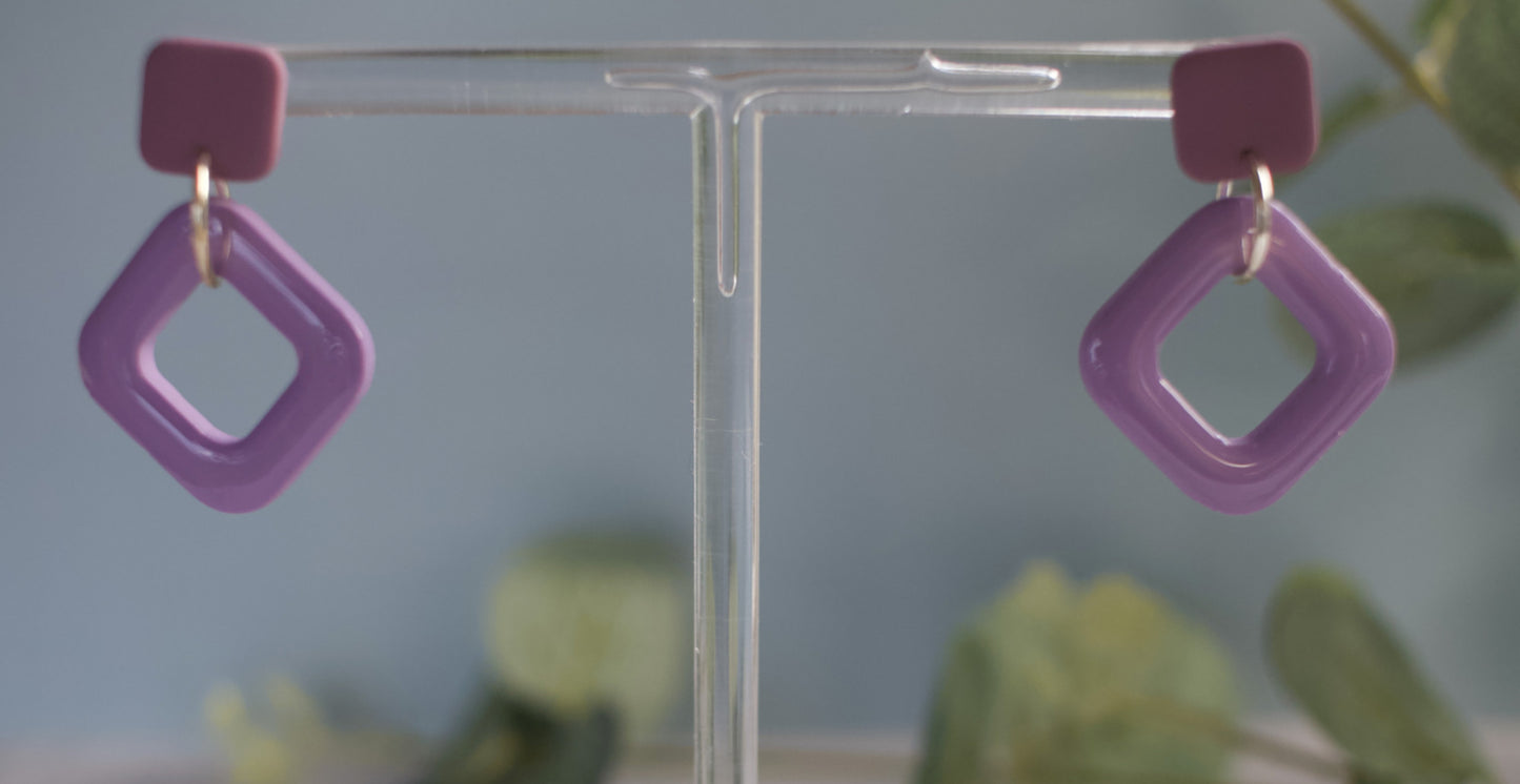 Purple square-shaped earrings on a clear stand with a blurred green background