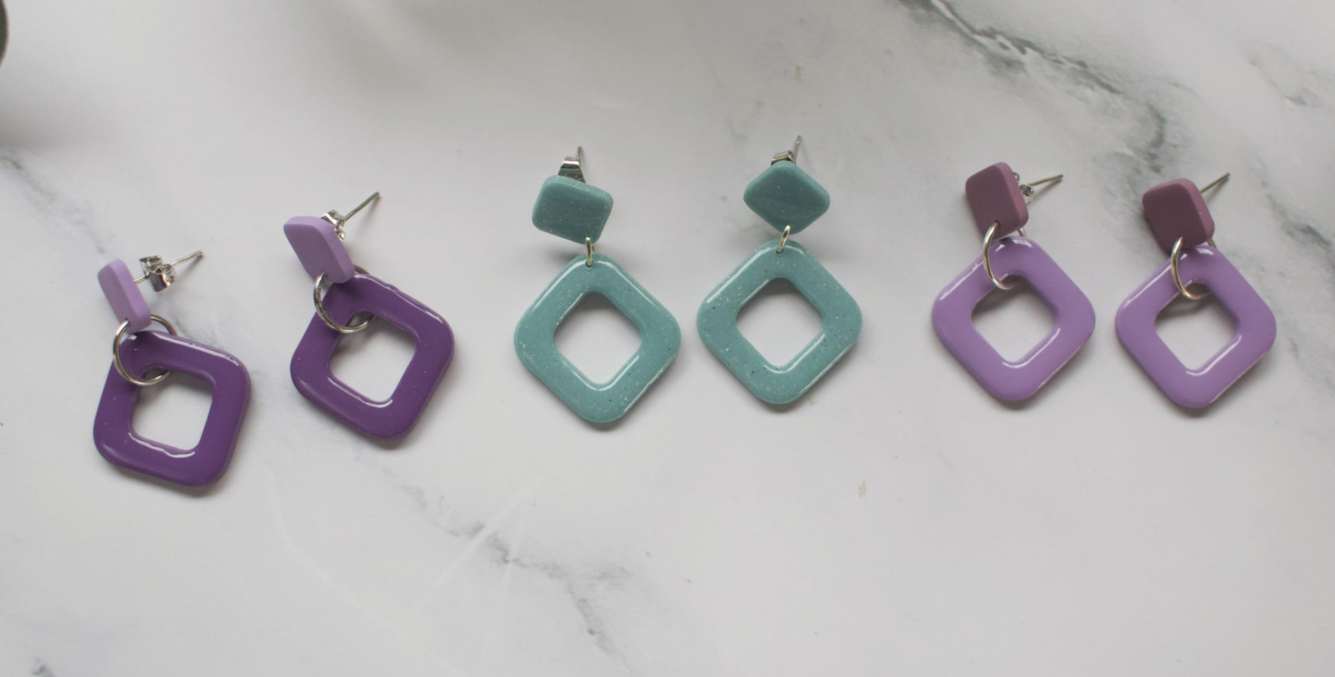 A collection of square-shaped earrings in purple and teal on a marble background