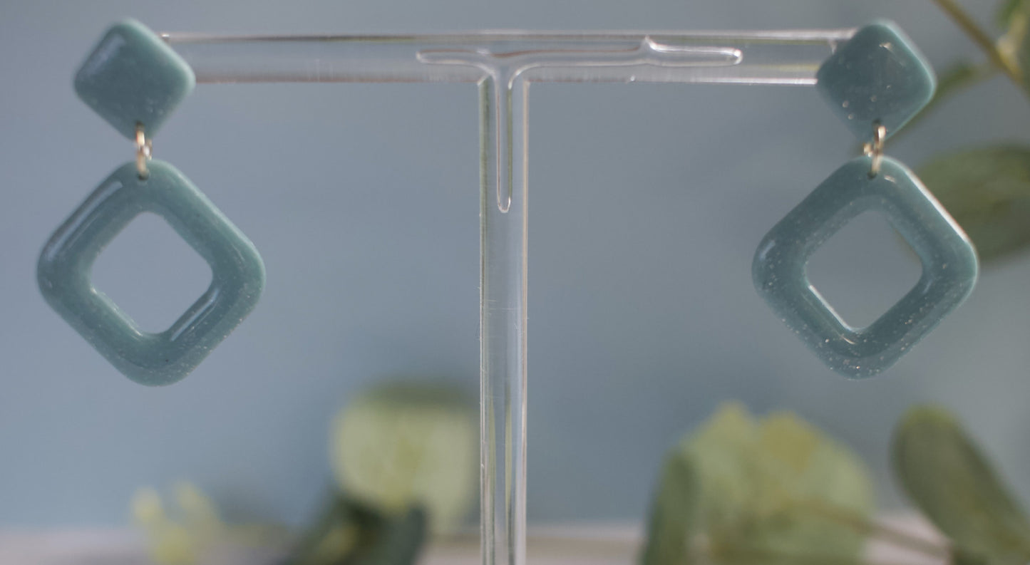 Teal square-shaped earrings on a clear stand with a blurred natural background
