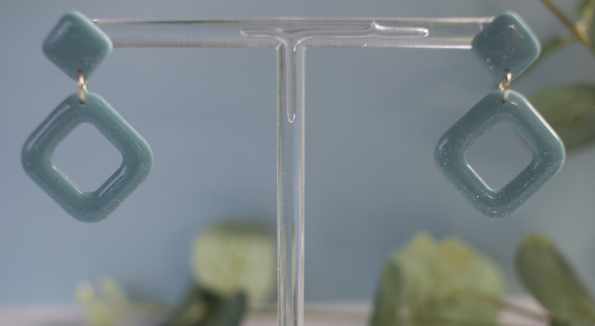 Teal square-shaped earrings on a clear stand with a blurred natural background