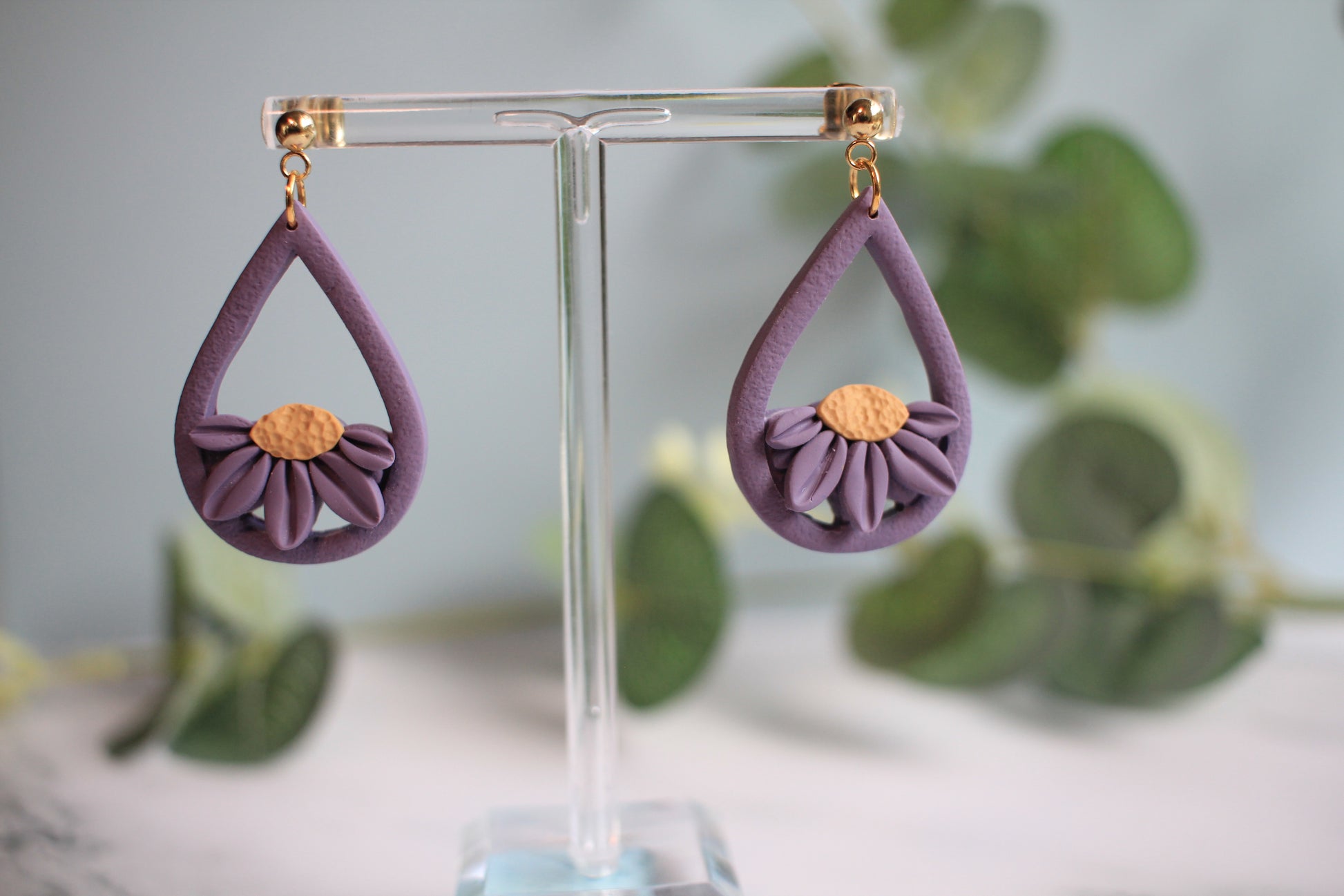 Purple teardrop earrings with floral design on a clear stand against a blurred greenery background
