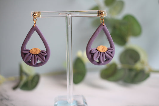 Purple teardrop earrings with floral design on a clear stand against a blurred greenery background
