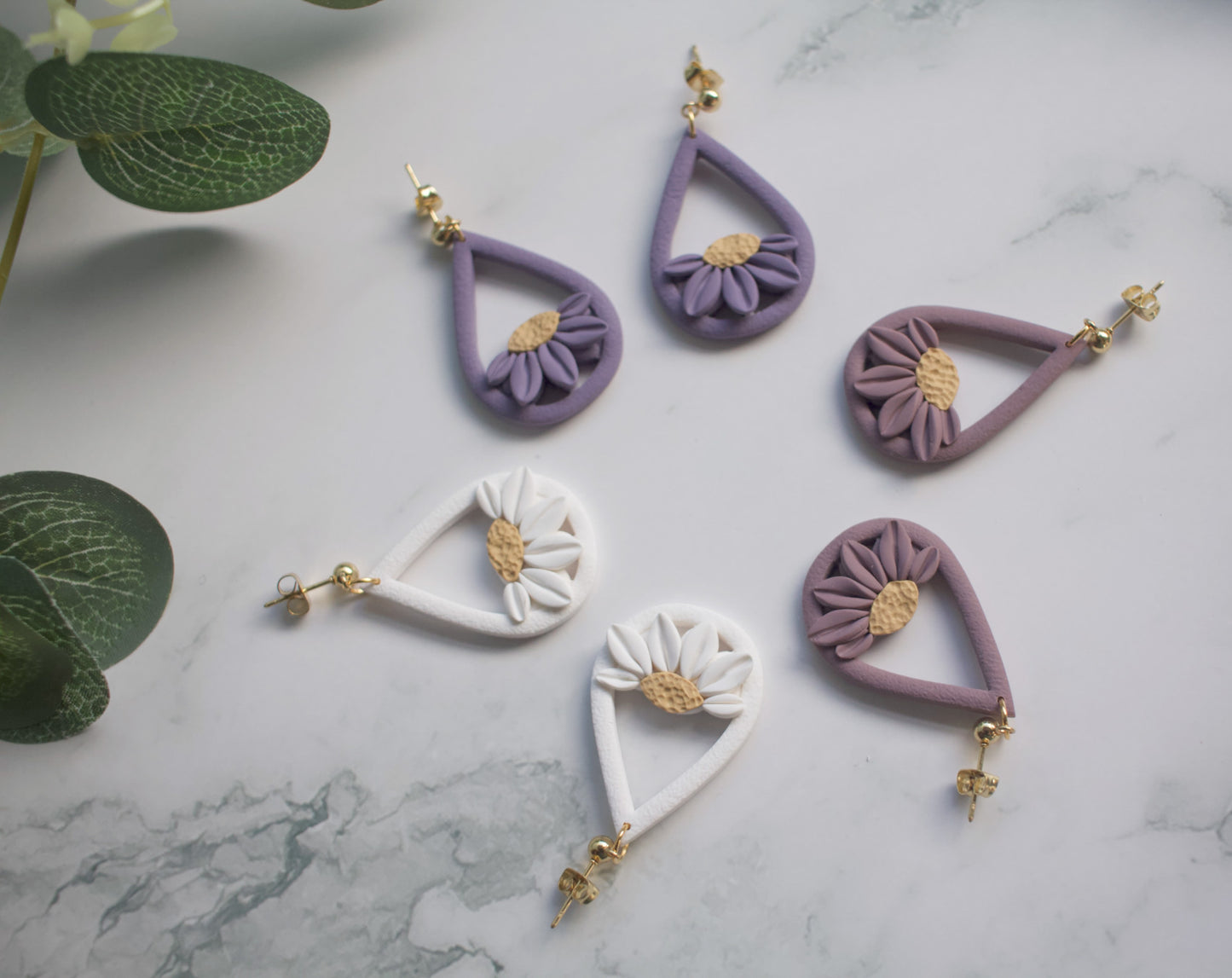 Collection of purple and white floral teardrop earrings on a marble surface with green leaves.